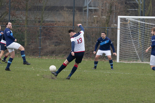 Youth soccer player in white and dark striped jersey kicks ball during match with blue-uniformed opponents and goal visible in background.