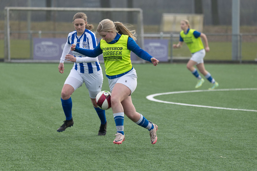 Female soccer player in yellow vest controls ball while being defended by opponent in blue and white striped jersey.