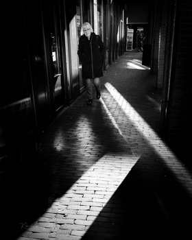 Person standing in a narrow alleyway with dramatic light and shadow creating geometric patterns on the wet pavement.