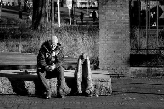 Bald man sitting on a wooden bench in black and white, looking downward with a contemplative expression.