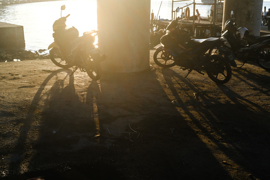 Motorcycles backlit by golden sunset, casting long shadows across dusty ground near industrial structures.