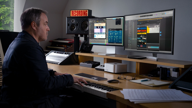 Music producer working at a studio desk with dual monitors displaying audio editing software and a keyboard controller.