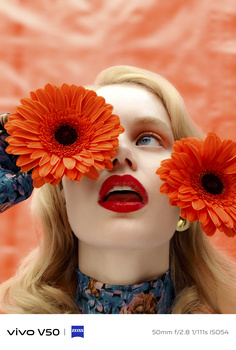 Woman with blonde hair and red lips partially obscured by two large orange gerbera daisies against a coral background.