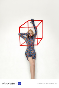 Woman posing against geometric red framework with arms raised, wearing patterned dress against white background.