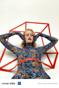 Woman in patterned blue and orange dress posing with arms behind head against geometric red frame backdrop.