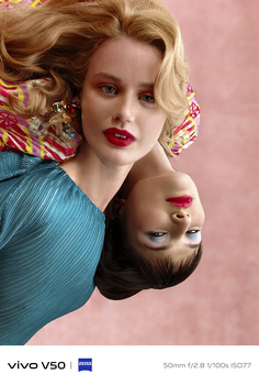Two women with styled blonde hair and bold red lipstick pose against a pink background in complementary blue garments.
