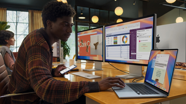 Man in plaid shirt working at wooden desk with laptop and dual monitors displaying data dashboards.