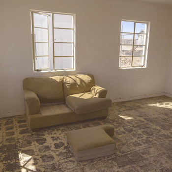 Minimalist interior with tan armchair and ottoman bathed in natural window light casting geometric shadows on textured floor.