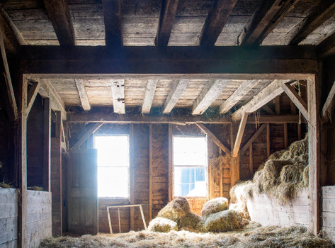 Rustic barn interior with hay piles and wooden beam structure, bright light streaming through two windows.