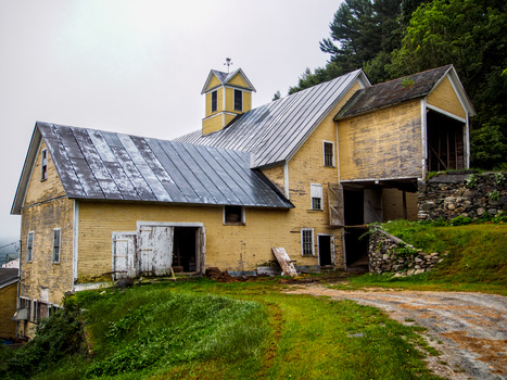 Abandoned yellow farmhouse with metal roof and stone foundation surrounded by overgrown grass.