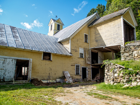 Abandoned farmhouse with yellow siding, stone foundation, and metal roof featuring solar panels.
