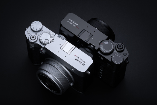 Two mirrorless cameras with lenses arranged on black surface, showing top-down view of controls and dials.