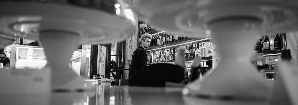 Black and white photograph of a person in a retail shop interior, viewed through glass shelving with out-of-focus foreground elements.