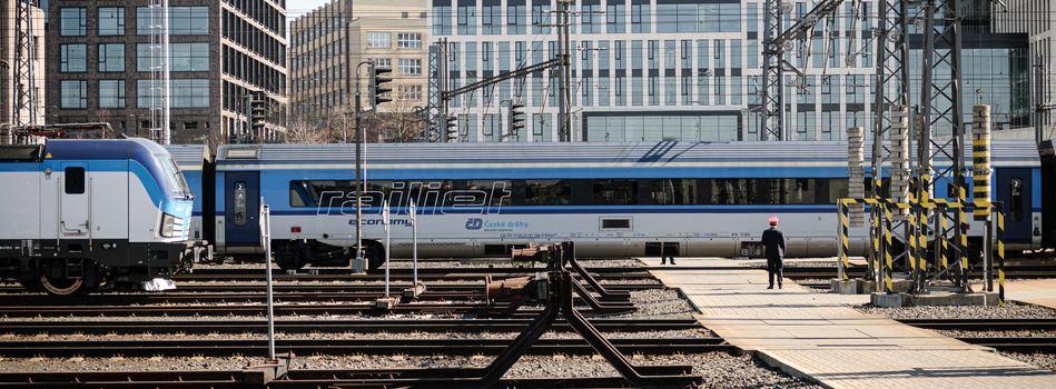 Blue and white commuter train parked at urban rail yard with downtown buildings and overhead power lines.