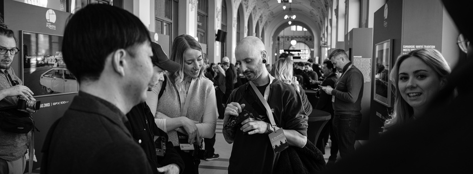 Black and white photograph of people conversing in a gallery corridor with arched ceilings.