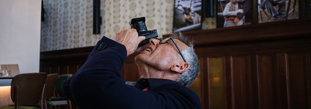 Man in navy blazer holding binoculars to his eyes while looking upward indoors.
