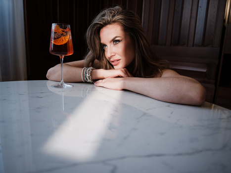 Woman in elegant pose at marble table with cocktail, soft window lighting creating dramatic shadows.