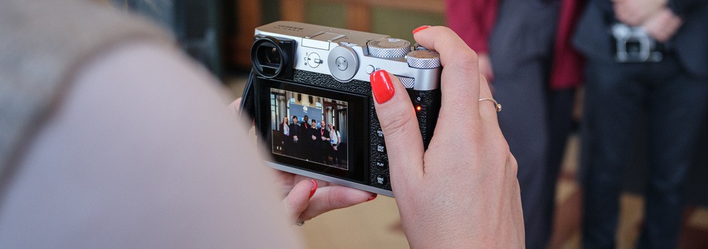Person holding a compact rangefinder camera displaying a live preview on its rear LCD screen.