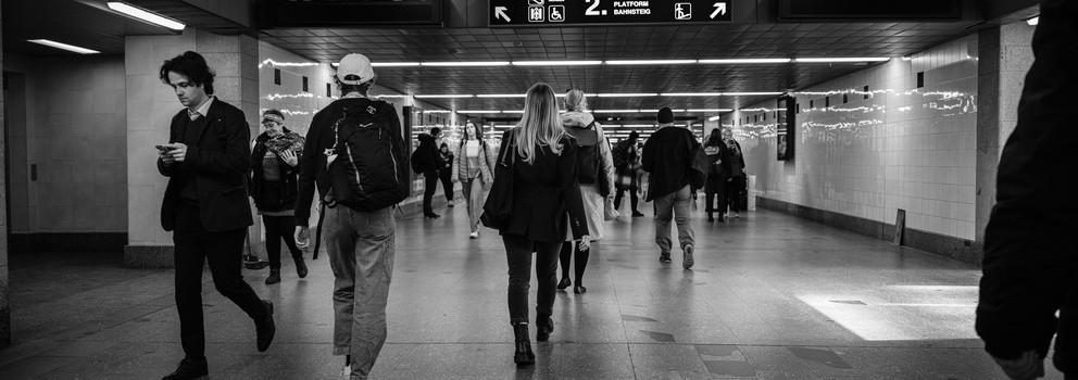 Commuters walking through a subway station corridor lit by fluorescent ceiling lights.