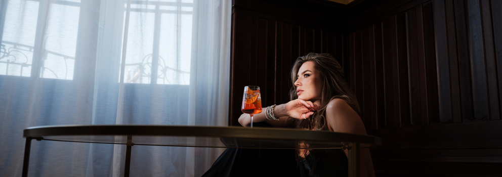 Woman at window holding drink, contemplative pose in moody interior lighting.