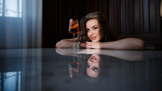 Woman leaning on reflective table with cocktail glass, dramatically lit with window light and dark background.