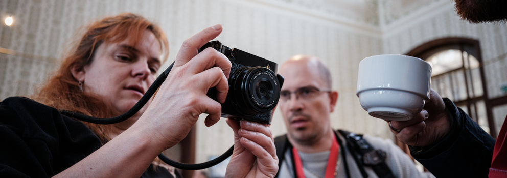 Woman holding a compact camera at a photography workshop or event, with attendees and architectural details visible in background.