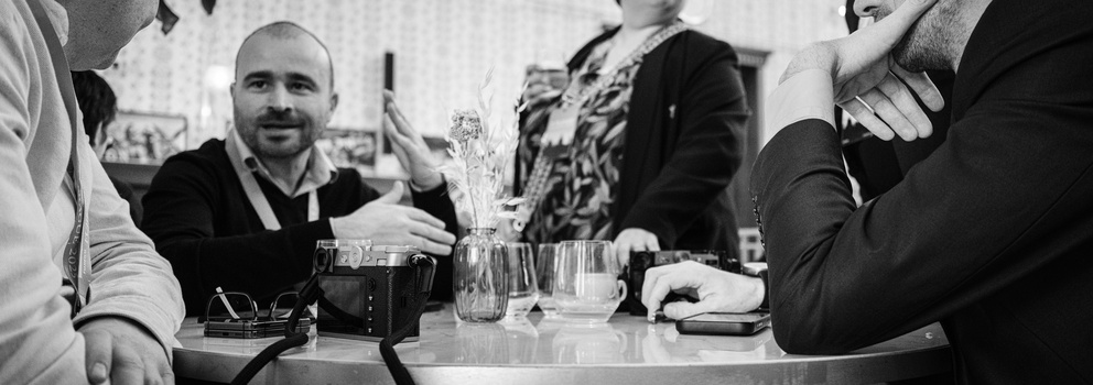 Man with mustache speaking at a table surrounded by people during what appears to be a professional meeting or event.
