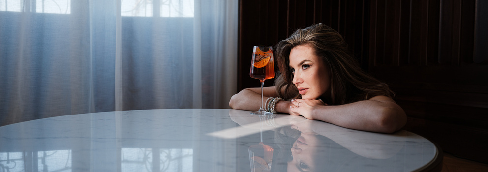 Woman leaning on a white table with a cocktail glass, backlit window in soft focus behind her.