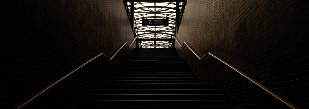 Symmetrical staircase ascending toward a skylight, shot from ground level with dramatic shadows and gold handrail lighting.