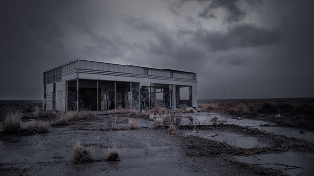 Abandoned concrete building with broken windows in a desolate, muddy landscape under stormy sky.
