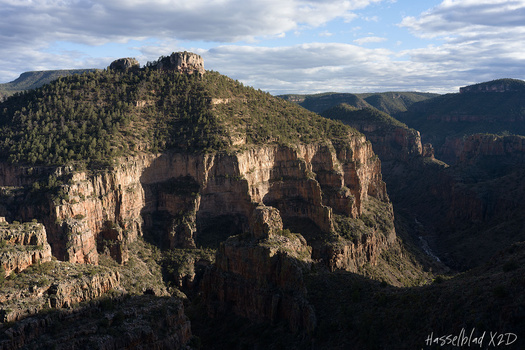 Dramatic rocky canyon landscape with towering cliff formations and forested ridgeline under partly cloudy sky.