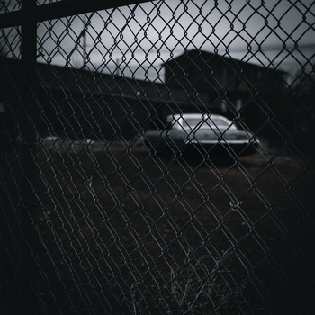 Silhouetted car visible through chain-link fence with industrial building in background.