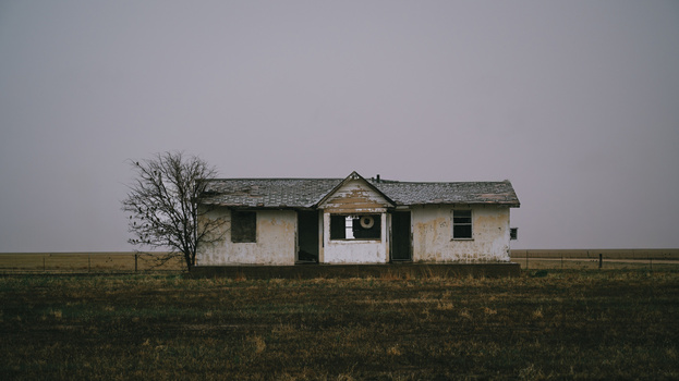 Abandoned single-story house with weathered white and tan exterior standing alone in a flat, barren landscape.