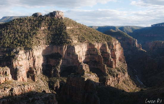 Steep rocky mountain peaks with forested slopes and deep canyon valleys under partly cloudy sky.