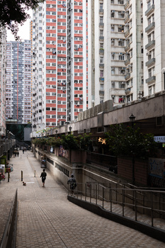 Urban street with tall residential buildings and pedestrians walking on a paved plaza beneath overcast sky.