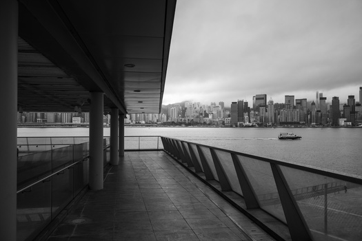 Urban waterfront promenade with metal railings overlooking a city skyline across the water.