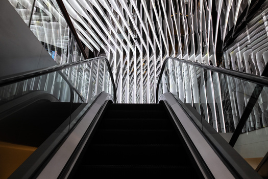 Looking up an escalator toward a modern building's ribbed ceiling with dramatic linear patterns and natural light.