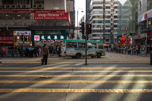 Busy Hong Kong street intersection with crosswalk, bus, and dense urban buildings in afternoon sunlight.
