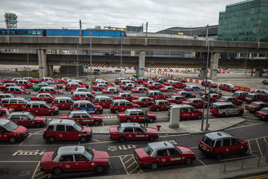 Overhead view of a large parking lot filled with dozens of red taxis arranged in organized rows beneath an elevated highway overpass.