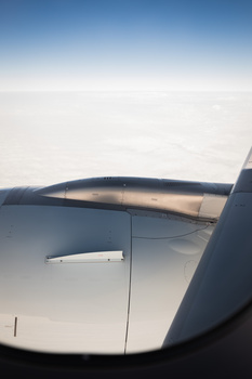 Aerial view from aircraft window showing wing and engine against cloudy sky and landscape below.