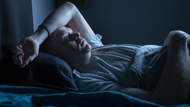 Man lying in bed at night holding a pillow over his face in a gesture of frustration or distress.
