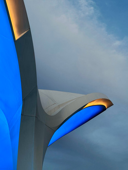 Modern architectural light fixture with glowing blue and orange elements against cloudy sky.