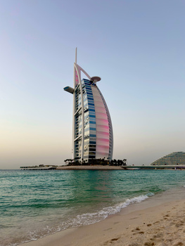 Sail-shaped luxury hotel tower with illuminated pink and blue facade rising from turquoise waters at sunset.