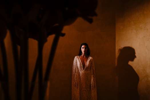 Woman in ornate robe standing under warm studio lighting with dramatic shadows cast on ochre wall.