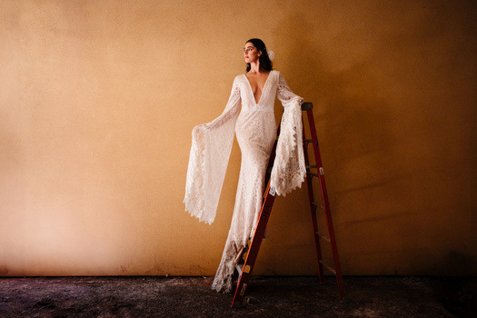 Woman in flowing white lace garment draped over wooden ladder against warm tan backdrop.
