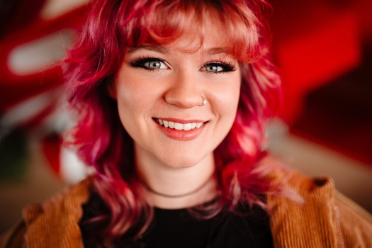 Woman with bright pink wavy hair and bangs smiling at camera against red bokeh background.