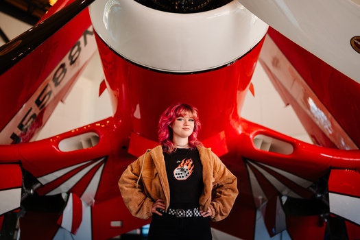 Person with pink hair standing confidently in front of a large red and white aircraft tail section.