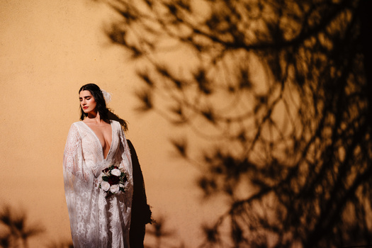 Bride in lace wedding dress holding bouquet with tree shadow cast on tan wall behind her.