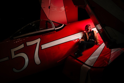 Person sitting inside a red aircraft fuselage marked with the number 57, illuminated by red studio lighting.