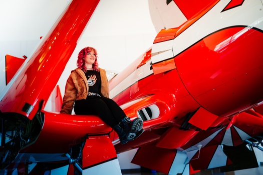 Person in brown jacket sitting on red aircraft fuselage against bright sky.